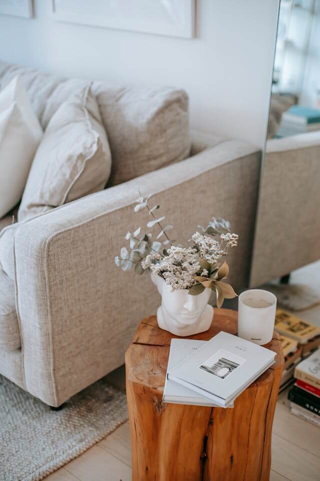 A cozy living room corner with a beige sofa, a wooden side table displaying a vase with flowers, a candle, and a book.