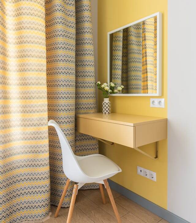 A minimalist vanity area with a yellow wall, patterned curtains, a wall-mounted mirror, and a chair.