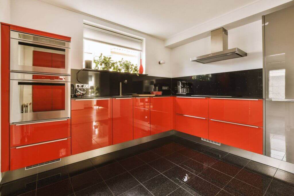 A modern kitchen with glossy red cabinets, a black countertop, stainless steel double ovens, a range hood, and a black tile floor. There are plants and a red vase on the windowsill.