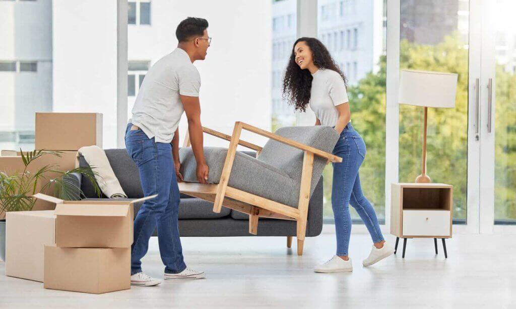 Two people are lifting a gray armchair in a room with moving boxes and modern furniture. Large windows reveal a city view with trees.