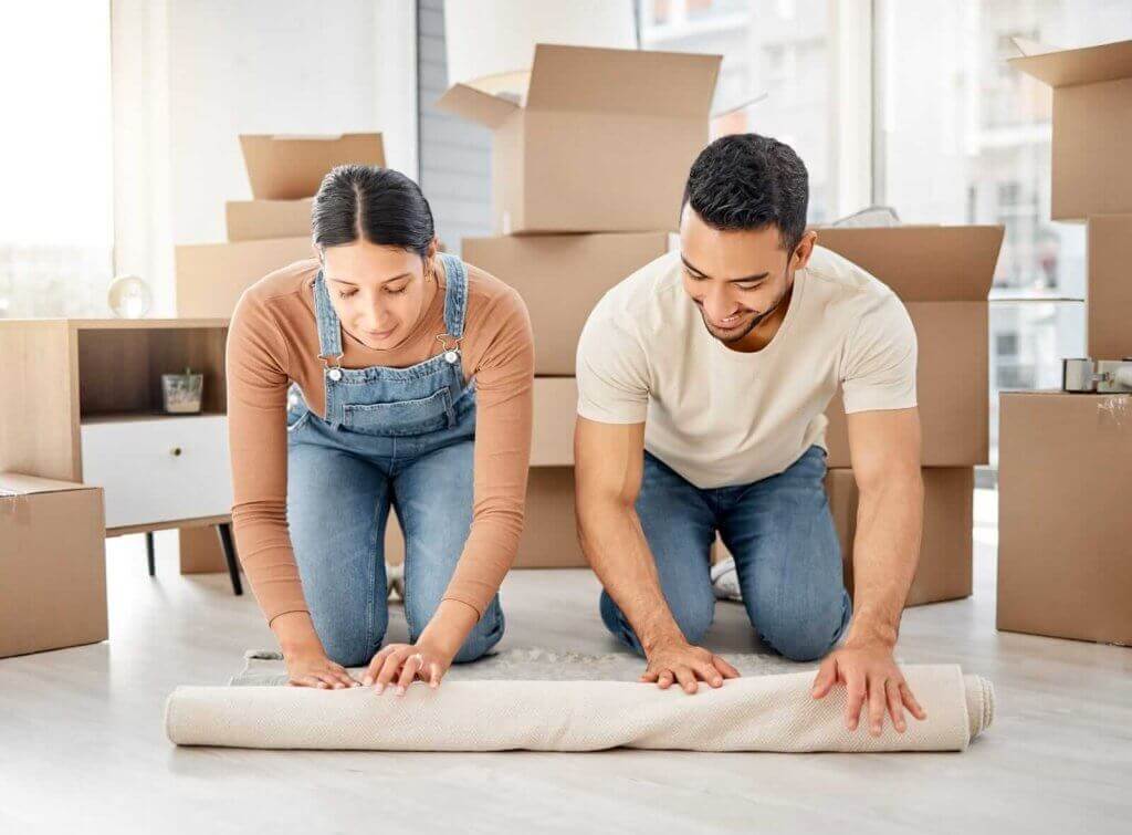 Two people kneeling on the floor together, rolling up a carpet in a room filled with cardboard moving boxes.