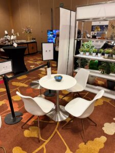 A Replica Tulip Table with a silver base and round white top is paired with three white chairs, featuring a central bowl and surrounded by potted plants on shelves in a carpeted display room.