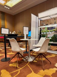 A Replica Tulip Table with a Silver & Round Top in White is paired with three white chairs on a patterned carpet in a conference room, featuring display screens and plants in the background.