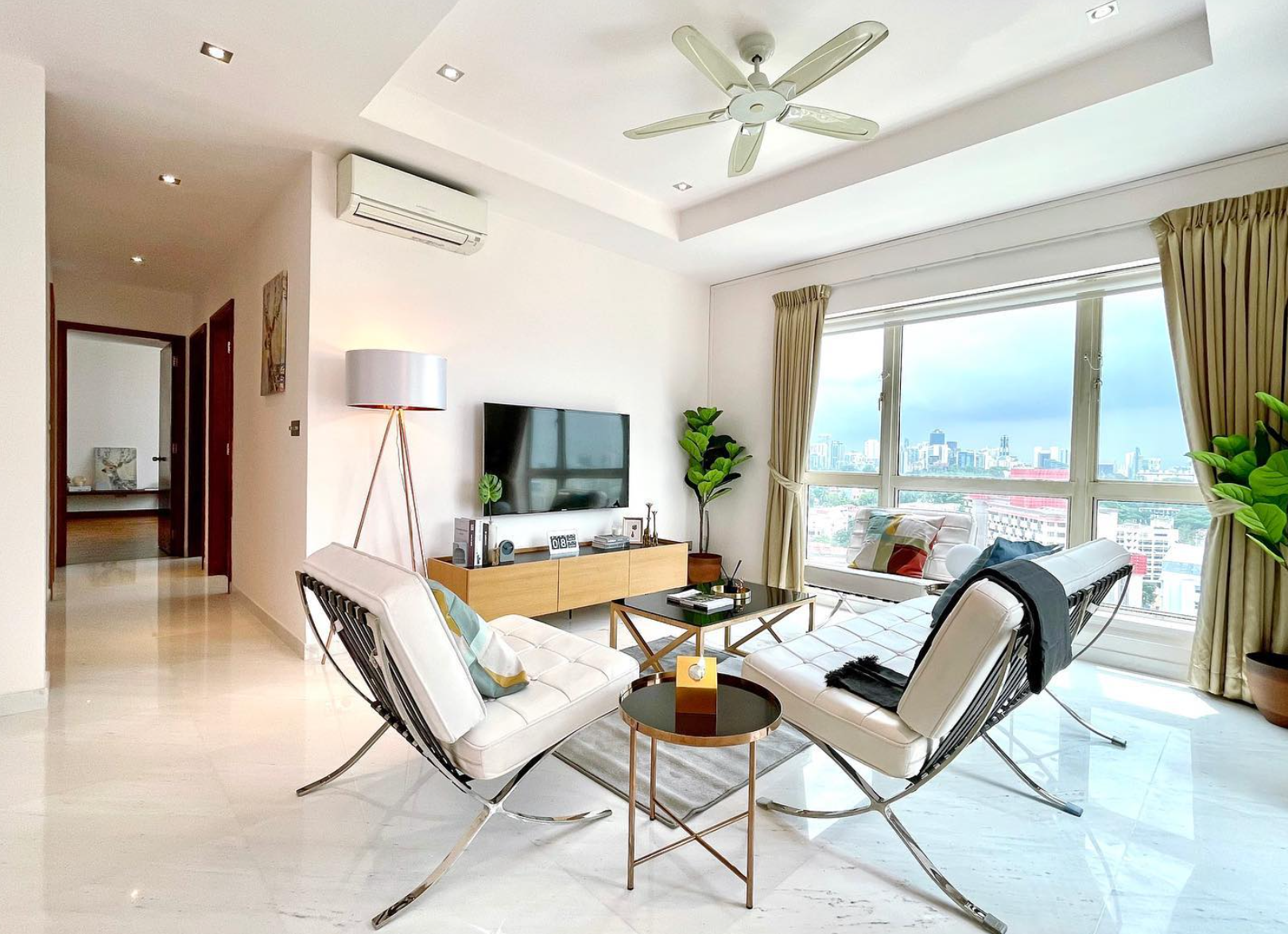 Modern living room with white furniture, marble floors, a flat-screen TV, large window with city view, and indoor plants. A ceiling fan and air conditioner are visible.
