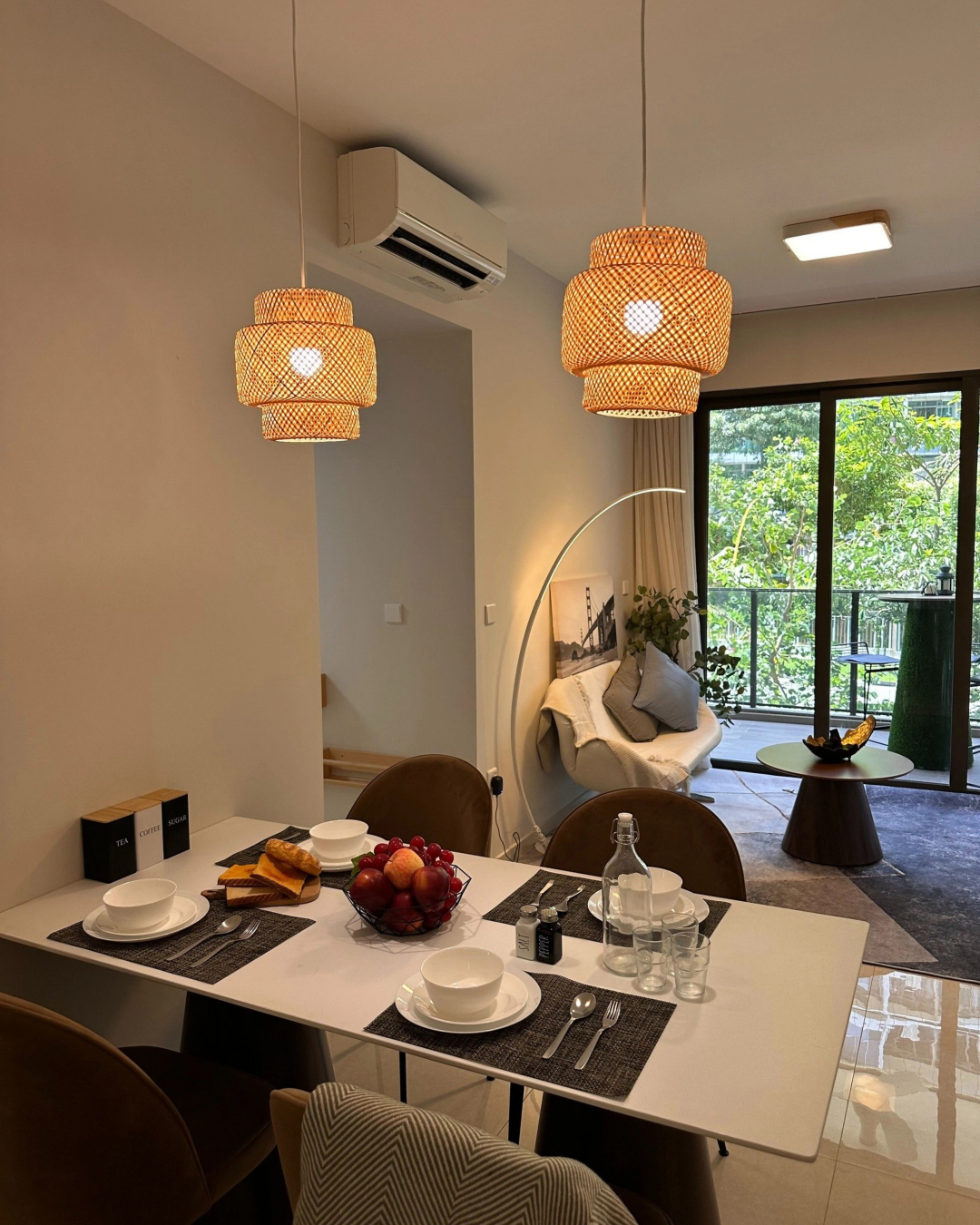 A modern dining and living area with a set dining table, pendant lights, a cozy chair by the window, fruit bowl, and large glass doors overlooking greenery.
