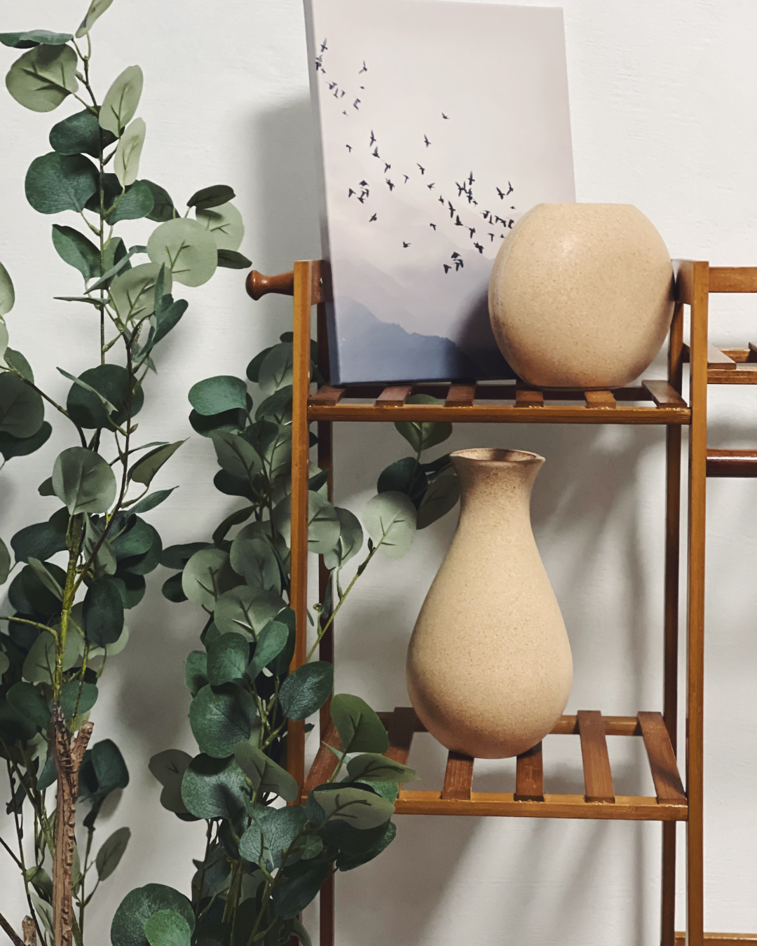 A wooden shelf holding two beige vases and a framed photo of birds in flight, with green leafy branches placed beside and around the shelf.