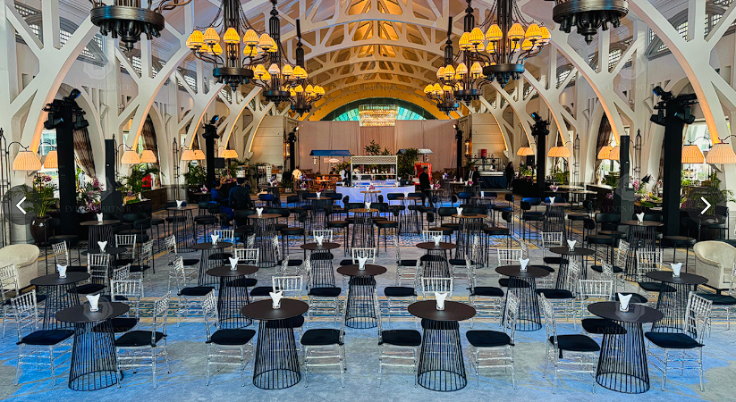 A spacious event hall with arched white beams, chandeliers, and rows of round tables with black chairs arranged facing a stage at the far end.