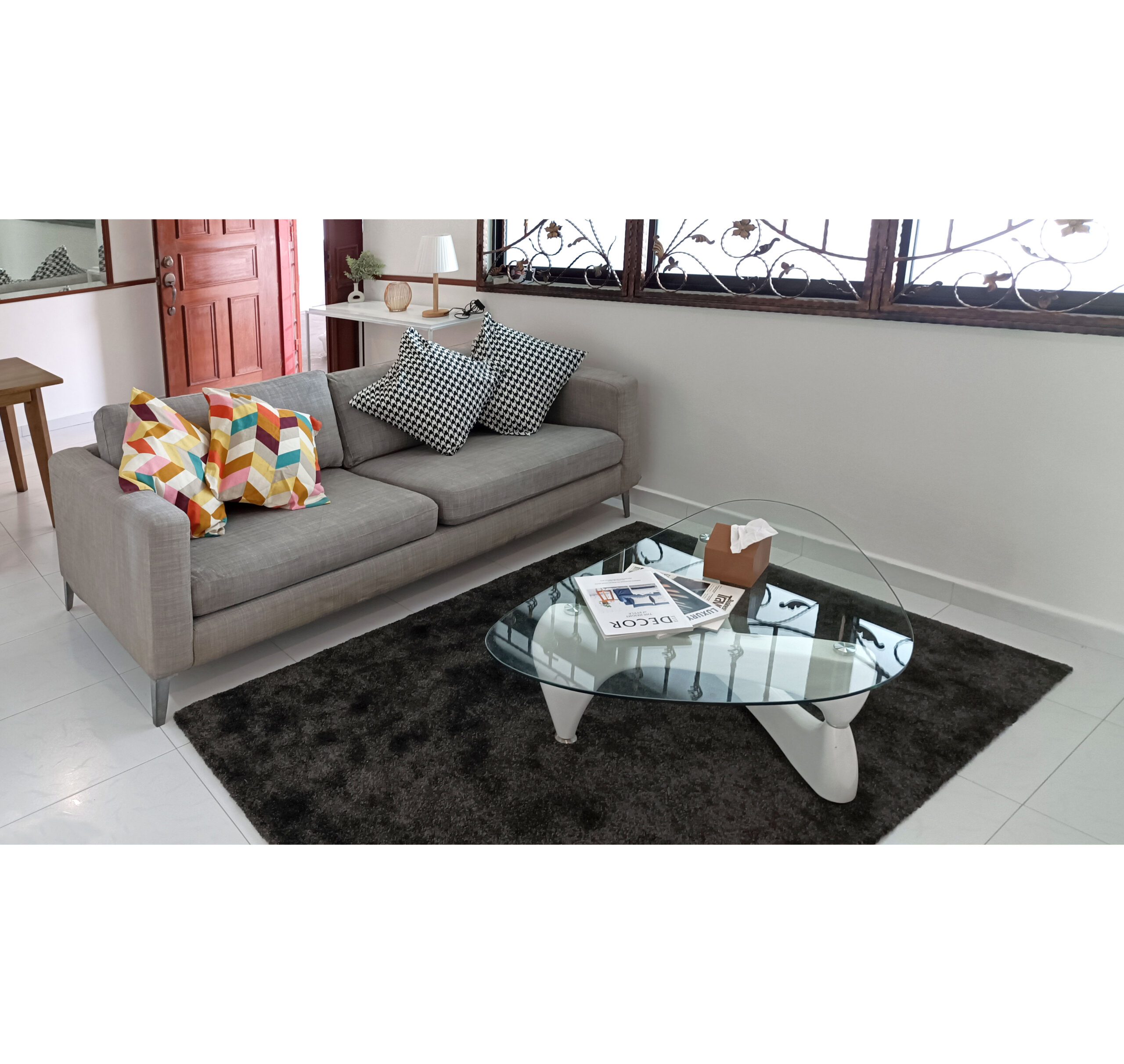 A Home Staging - Housing Development Board (HDB) flat features a modern living room with a gray sofa, patterned cushions, glass coffee table with books, and dark rug on a white tile floor.