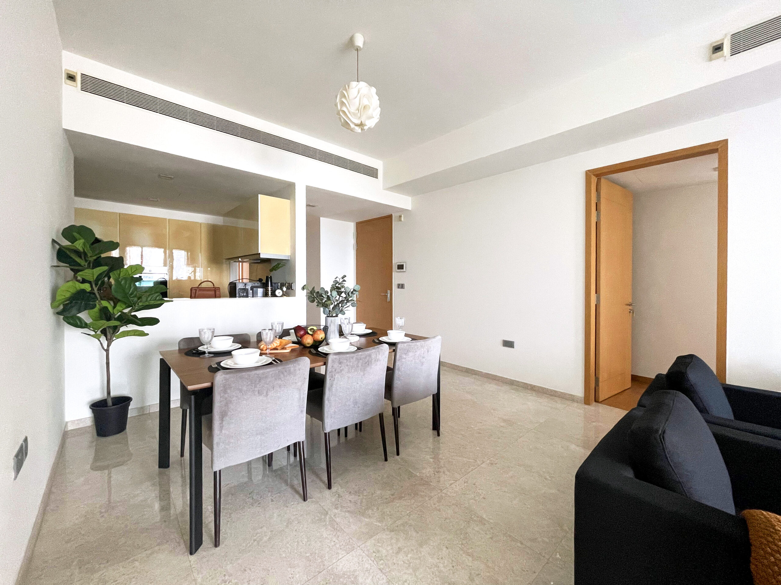A modern dining area with a table set for six, adjacent to an open kitchen, potted plant, and beige tiled floor in a bright, minimalist apartment.