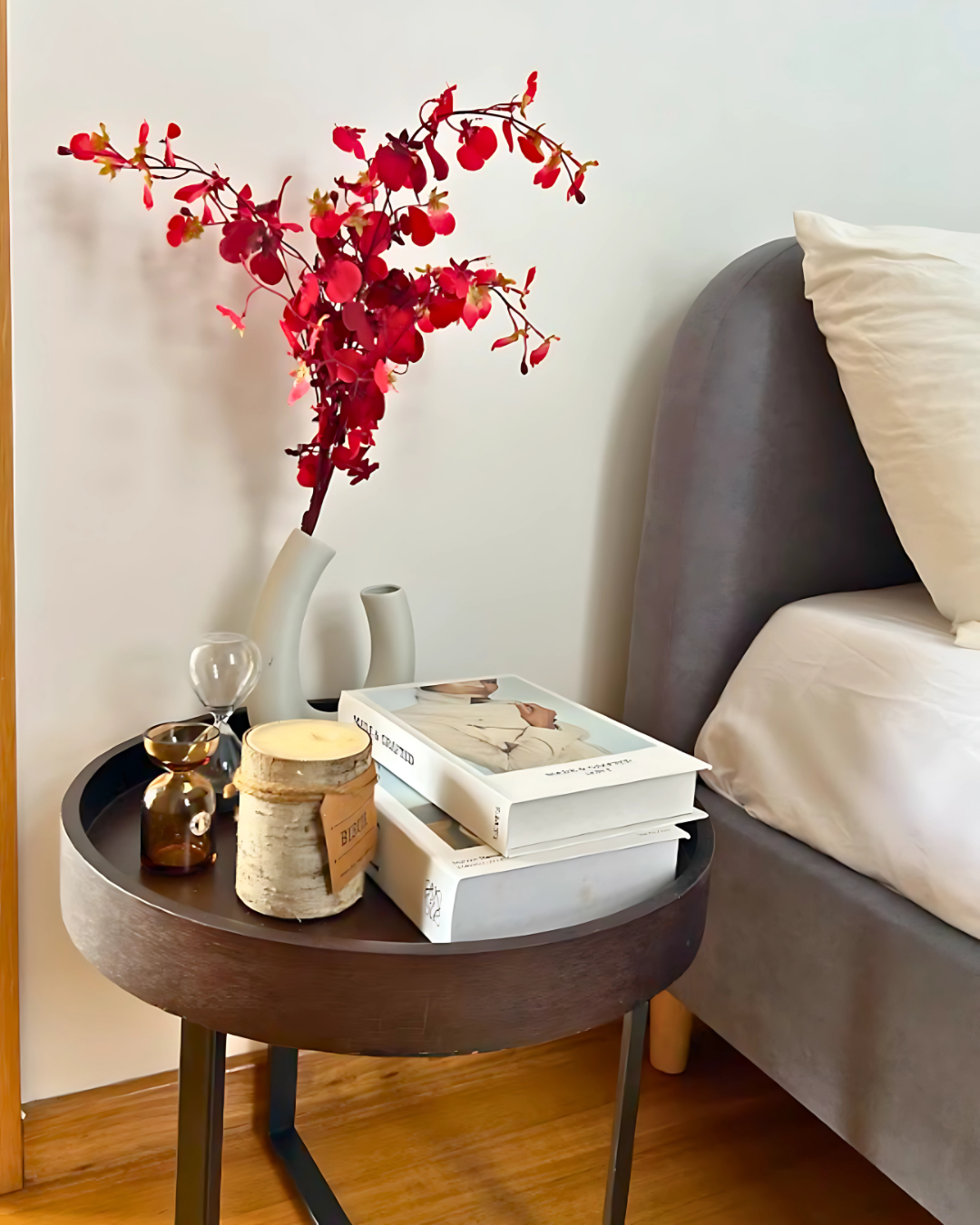 A round bedside table with stacked books, a white vase with red flowers, an hourglass, a candle, and a small jar next to a bed with white bedding.